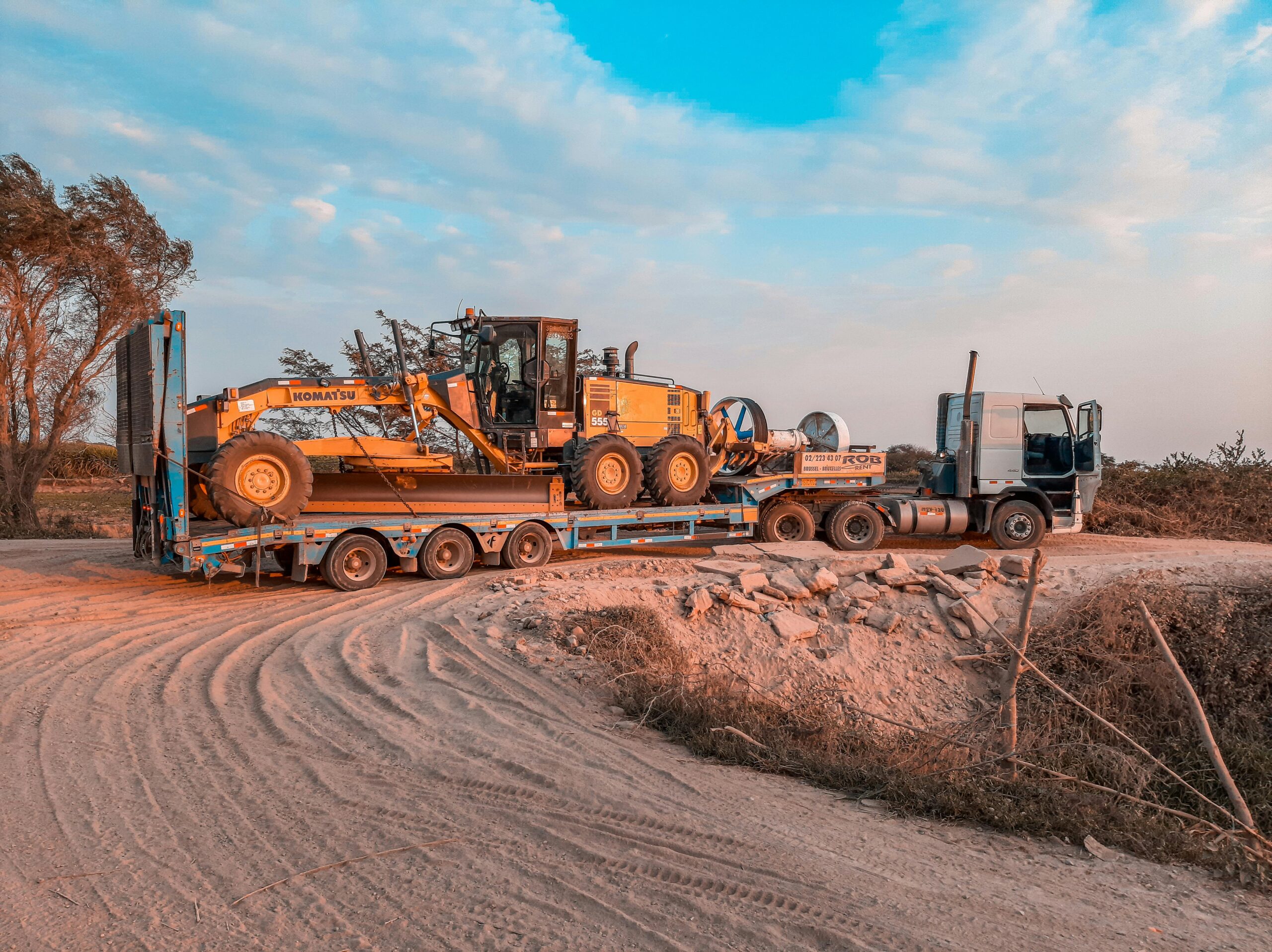 A cargo truck transporting heavy machinery on a rugged dirt road under a clear sky.